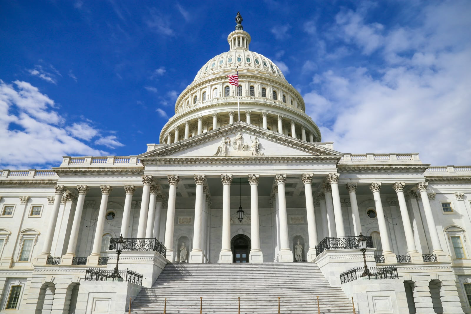 Low-angle view of the United States Capitol dome in Washington, D.C., showing white neoclassical columns, the grand staircase, and an American flag flying against a blue sky.