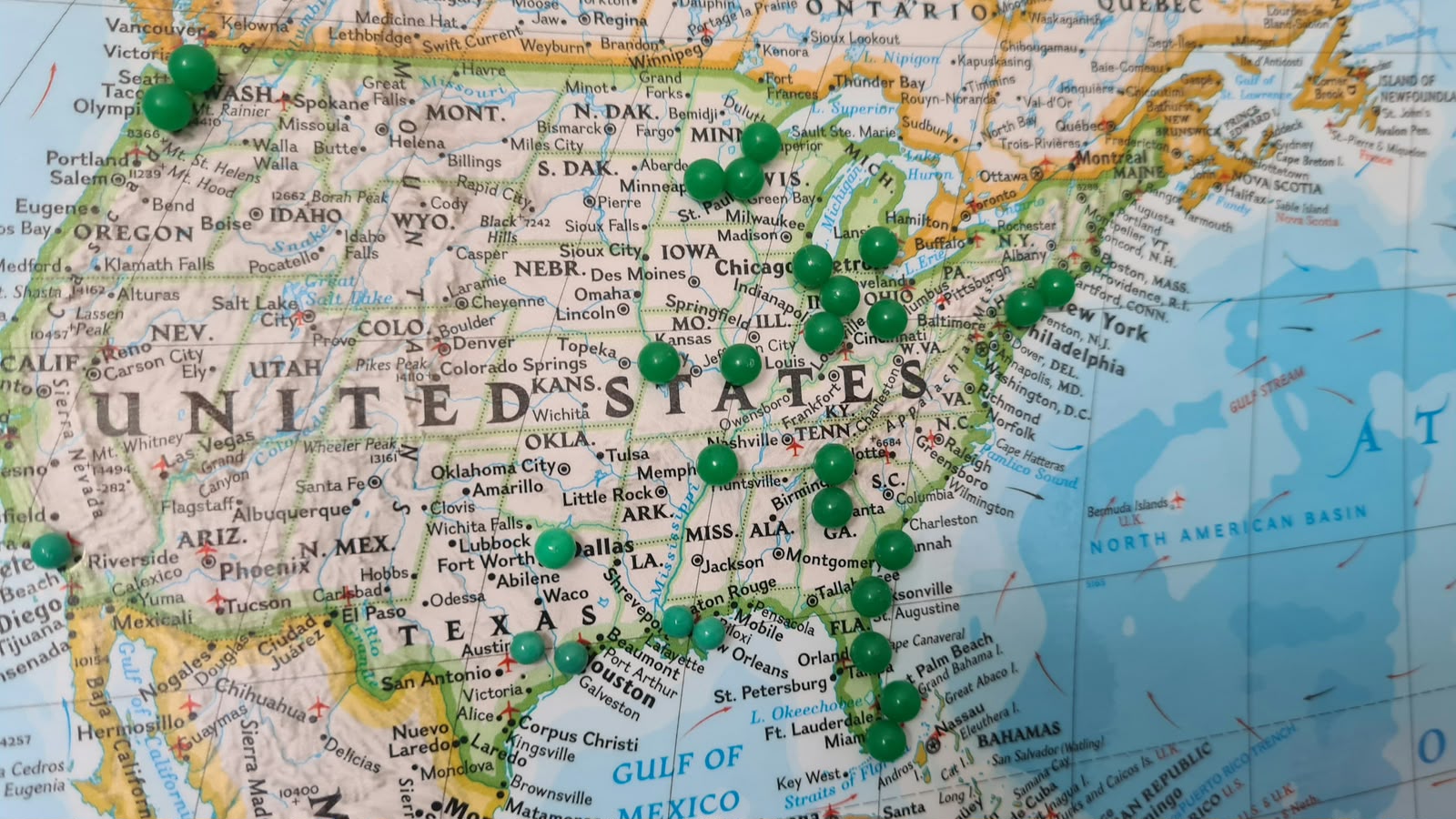 Close-up of a printed map of the United States with dozens of green pushpins placed across various cities and states.